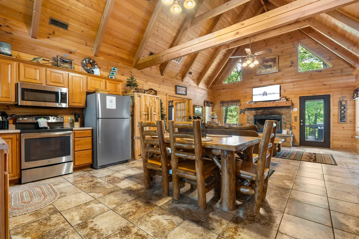 A wide-angle view of the open-plan dining area and kitchen, featuring a rustic log dining table, ample kitchen cabinetry, a view of the stone fireplace from another angle, and large windows revealing woodland scenery. The layout emphasizes togetherness and flow between cooking, dining, and relaxing spaces.