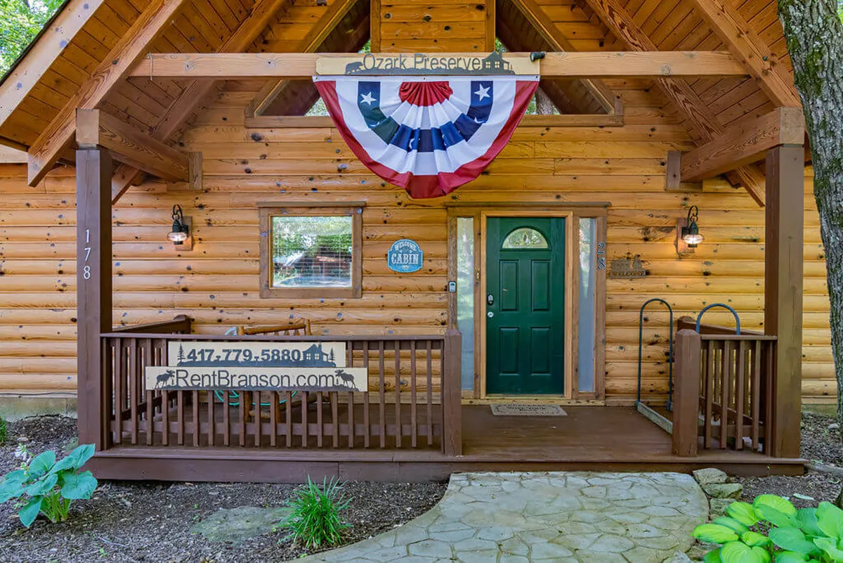A welcoming log cabin exterior featuring a green front door adorned with a decorative wreath, a cozy porch area with signage, and lush greenery surrounding the entrance. The scene suggests a place where guests can relax outdoors and soak up the festive, lively atmosphere of the area.