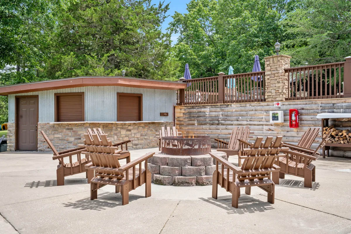 A cozy fire pit area surrounded by classic Adirondack chairs and a rustic stone wall, nestled among mature trees. The setting suggests a communal gathering spot ideal for evening s’mores, storytelling, and stargazing.