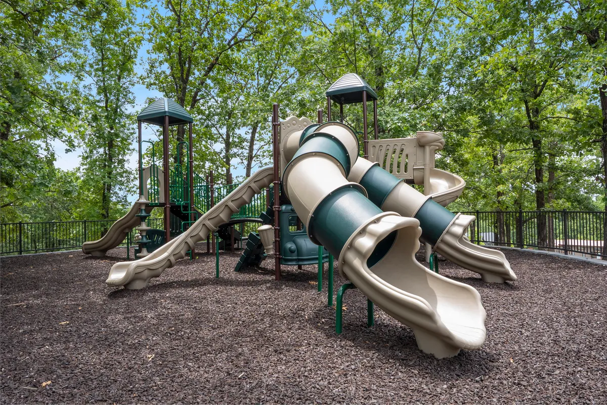 A colorful, well-equipped playground with slides and climbing structures, framed by tall trees and open space. Children can be seen playing, and there is room for families to gather and relax nearby.