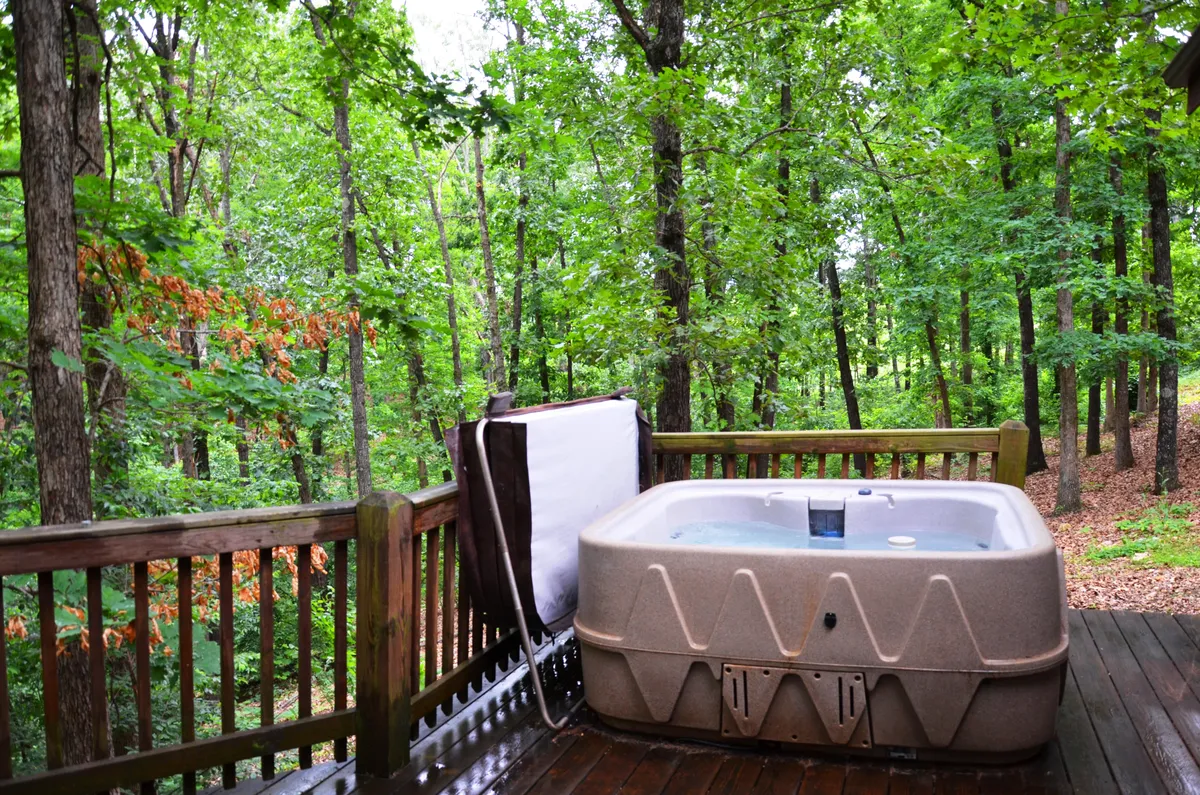 A close view of the hot tub on the deck, surrounded by towering trees and natural greenery. The serene scene hints at starlit soaks and peaceful moments spent immersed in nature, with the deck’s wood tones blending seamlessly with the forest backdrop.