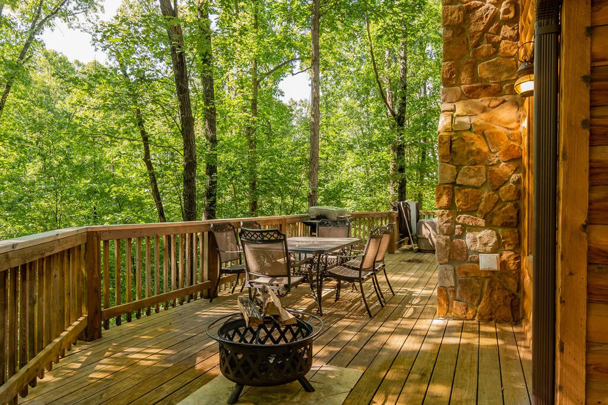 A serene wooden deck nestled among tall trees, featuring an outdoor dining set and a cozy fire pit. The area is surrounded by tranquil woods, creating a private, intimate atmosphere ideal for relaxing or enjoying meals outdoors.