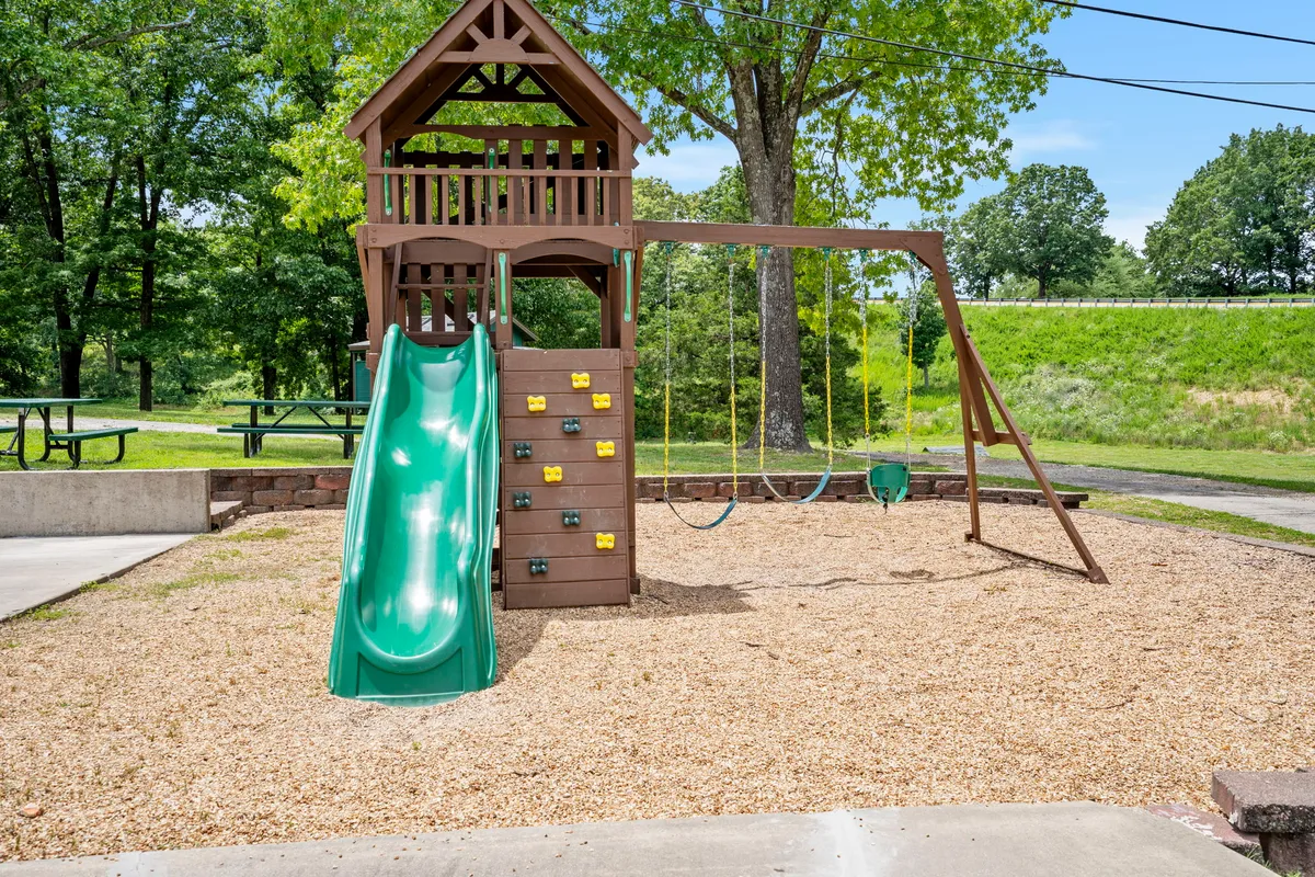 A dynamic shot of a playground structure with a slide, swing set, and climbing wall, set against a backdrop of green trees. Children are actively playing, capturing the joyful and energetic atmosphere that family travelers seek.