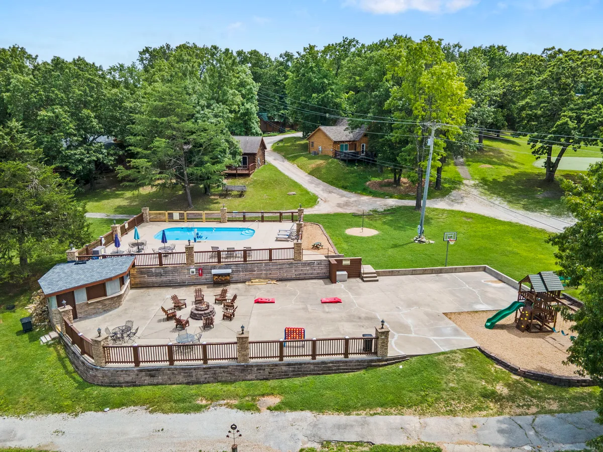A vibrant scene featuring a large swimming pool with lounge chairs and umbrellas, an outdoor seating area, a modern playground with slides and climbing structures, and a basketball hoop in view. Lush greenery and cabins surround the space, creating an inviting and communal resort atmosphere.
