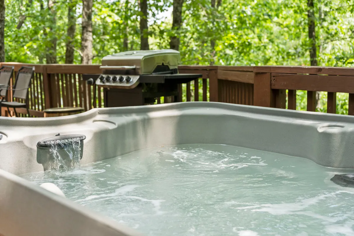 A peaceful exterior shot of a wooden deck nestled beside the forest, featuring a sparkling hot tub and a barbecue grill. The deck overlooks dense woods, creating a private escape ideal for soaking, grilling, and enjoying nature.
