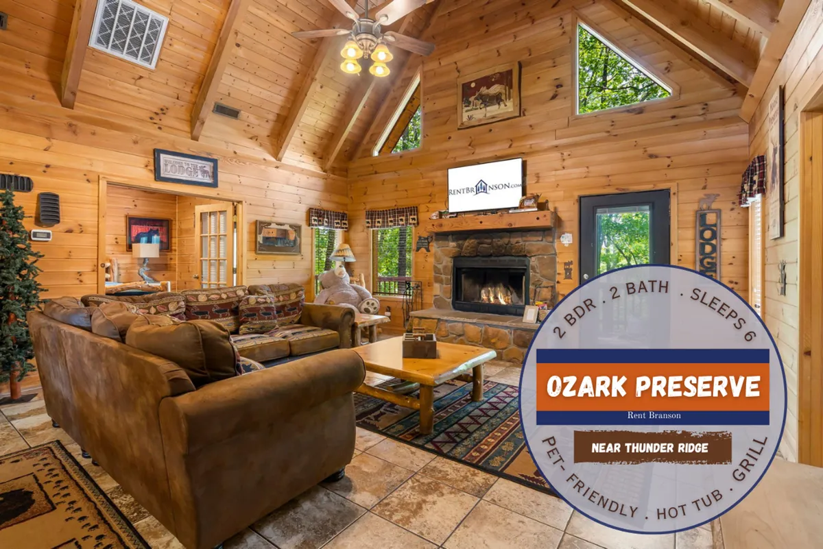 This interior image features a spacious great room with a large stone fireplace as the focal point. Cozy seating is arranged to face both the fireplace and a flatscreen TV, while wooden beams overhead and warm lighting create a rustic yet inviting atmosphere—ideal for group gatherings, movie nights, or relaxing conversations.