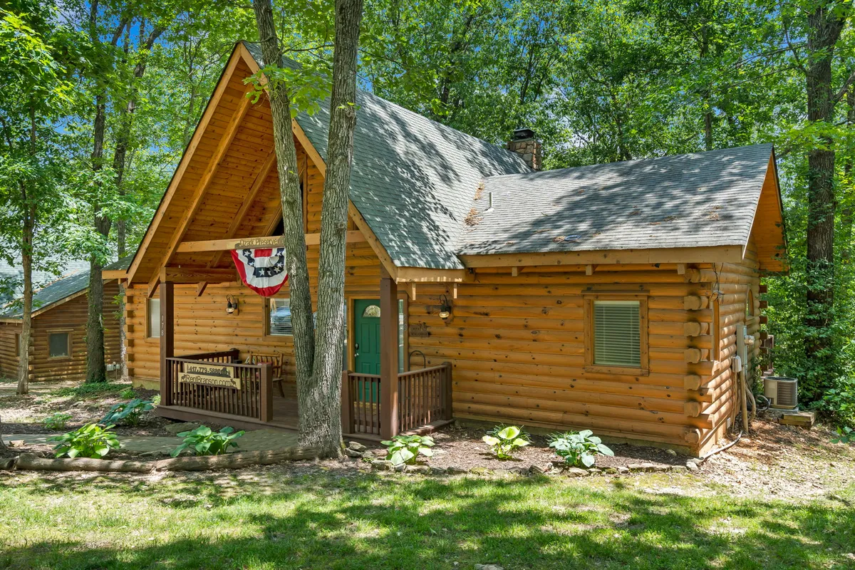 A welcoming log cabin exterior with a decorative banner and a cozy front porch area surrounded by lush natural greenery. The porch is furnished, inviting guests to relax and enjoy the fresh Ozark air. The setting emphasizes harmony with nature and a peaceful, eco-friendly atmosphere.
