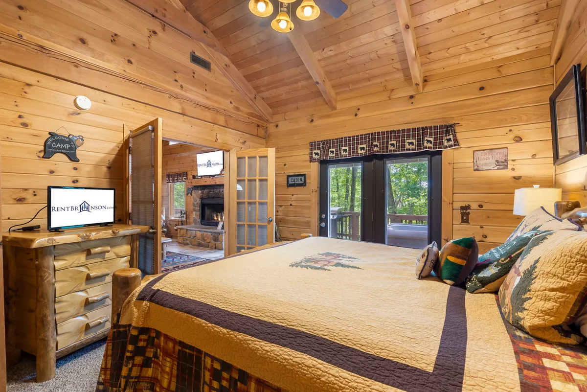 A cozy bedroom with a bed dressed in a patterned quilt, a wooden dresser, TV, and expansive windows that open up to views of nature. The natural light and tranquil setting evoke a sense of comfort and peaceful seclusion.
