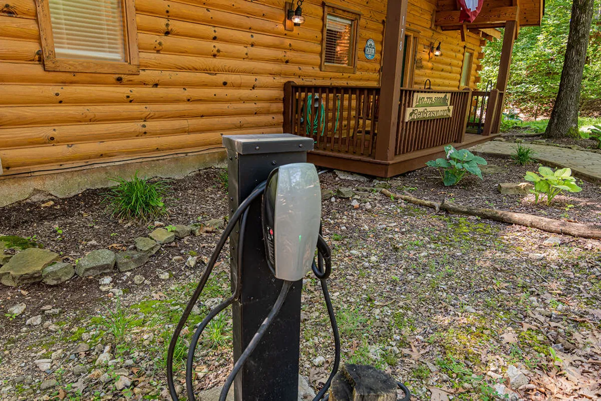 A welcoming exterior shot of a cabin fronted by a porch, with an electric vehicle charging station visible nearby and lush greenery surrounding the property. The scene feels modern, accessible, and eco-conscious.