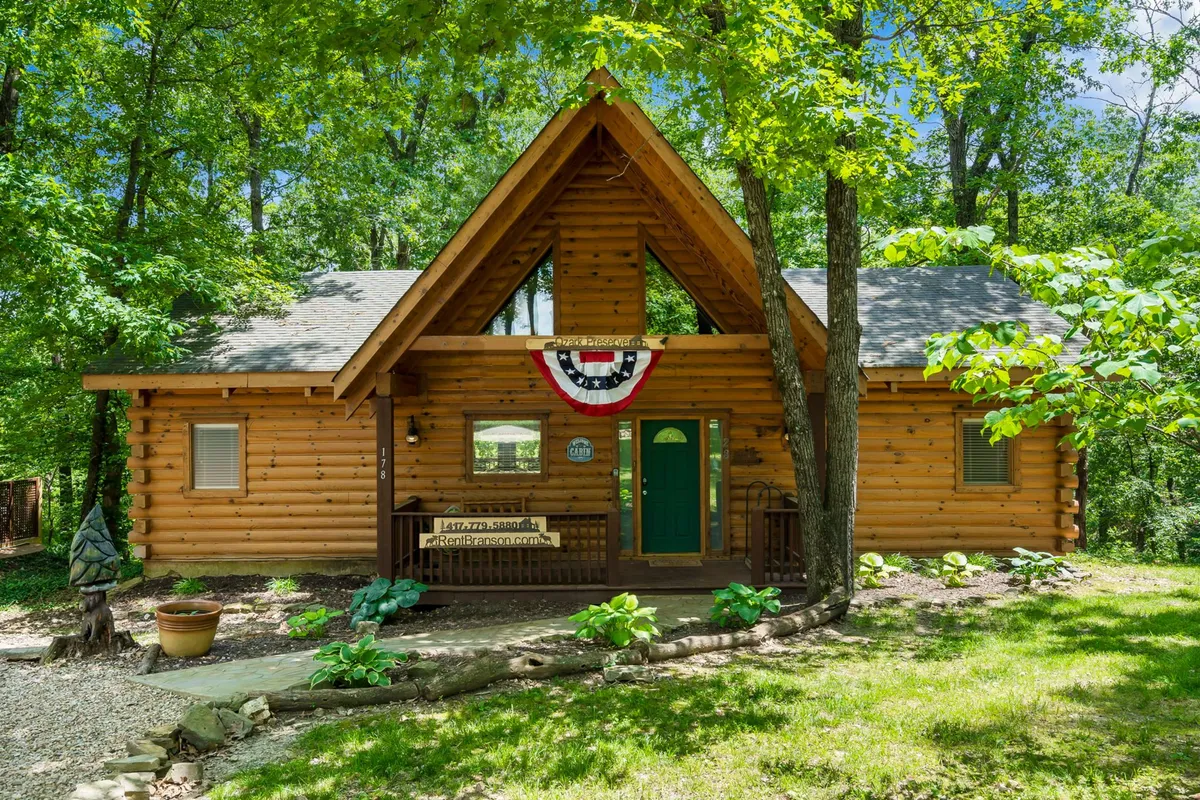 A cozy log cabin with a green door and decorative bunting sits nestled amid abundant greenery. The surrounding area hints at nearby trails and forested expanses, with a front porch ideal for relaxing after outdoor adventures.