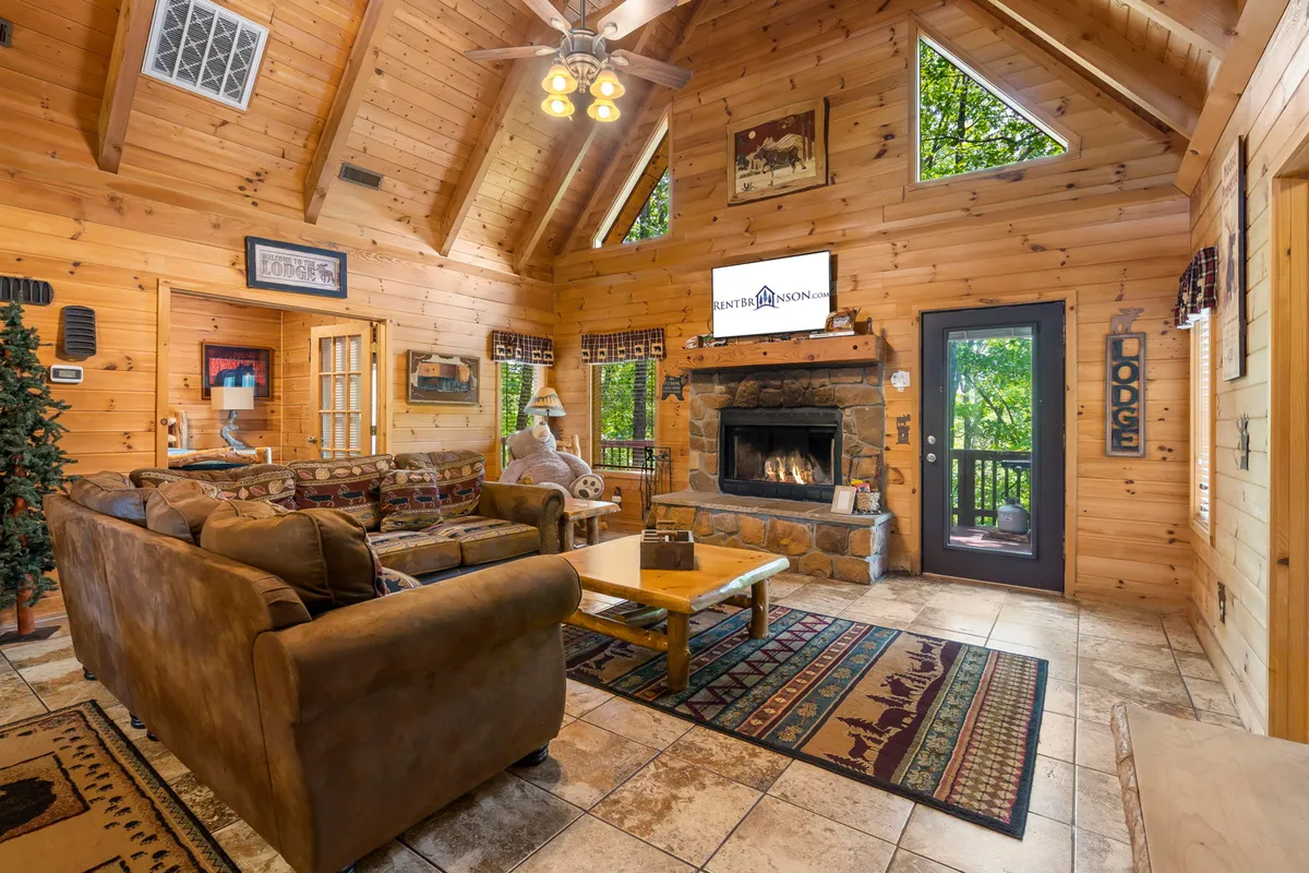 A cozy living room interior featuring a glowing stone fireplace, plush sofa, exposed wooden beams, and expansive windows framing views of the surrounding woods. The scene invites relaxation, warmth, and togetherness, perfect for unwinding after a day exploring the Ozarks.